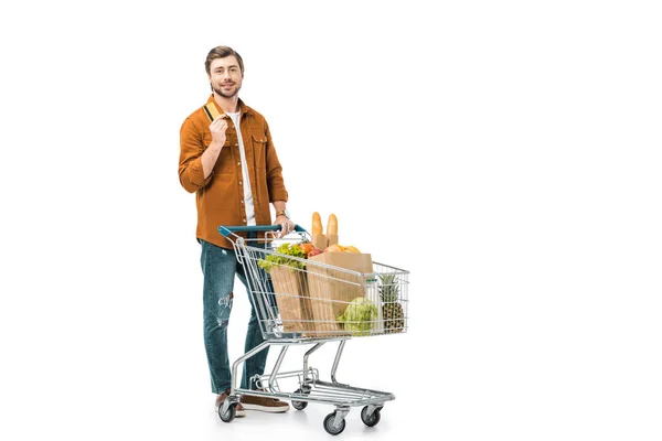 Homem que mostra o cartão de crédito ao estar perto do carrinho de compras com sacos de papel isolados no branco — Fotografia de Stock