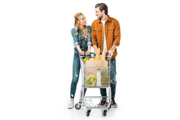 Casal feliz carregando carrinho de compras com produtos e olhando um para o outro isolado no branco — Fotografia de Stock