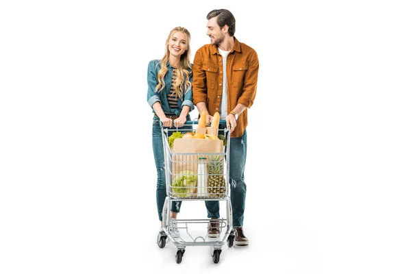 Усміхнена молода пара, що носить торговий візок з продуктами ізольовані на білому — стокове фото
