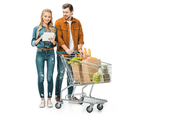 Attraktive lächelnde Frau checkt Einkaufsliste auf digitalem Tablet, während ihr Freund in der Nähe mit einem Einkaufswagen voller Papiertüten mit Produkten auf weißem Papier steht — Stockfoto