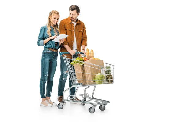 Hermosa mujer joven revisando la lista de compras en la tableta digital, mientras que su novio de pie cerca con el carro lleno de bolsas de papel con productos aislados en blanco - foto de stock