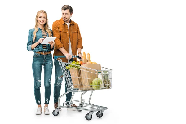 Mujer joven revisando la lista de compras en la tableta digital, mientras que su novio de pie cerca con el carro lleno de bolsas de papel con productos aislados en blanco - foto de stock