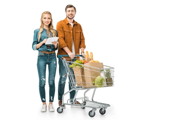 Chica feliz comprobando la lista de compras en la tableta digital, mientras que su novio de pie cerca con el carro lleno de bolsas de papel con productos aislados en blanco - foto de stock