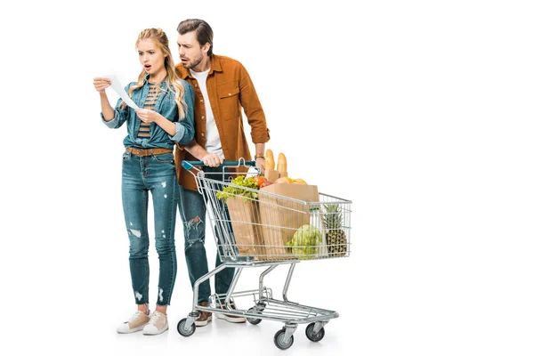 Casal chocado com carrinho cheio de produtos em sacos de papel olhando para verificação de compras isolado no branco — Fotografia de Stock