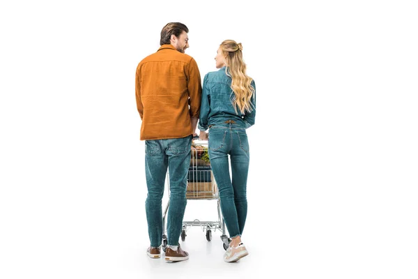 Vista trasera de pareja llevando carrito de compras con productos y mirándose aislados en blanco - foto de stock