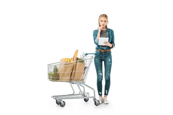 Donna premurosa guardando tablet digitale e in piedi vicino al carrello della spesa con prodotti isolati su bianco — Foto stock