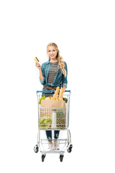 Junge Frau zeigt Kreditkarte und trägt Einkaufswagen mit Produkten auf weißem Grund — Stockfoto