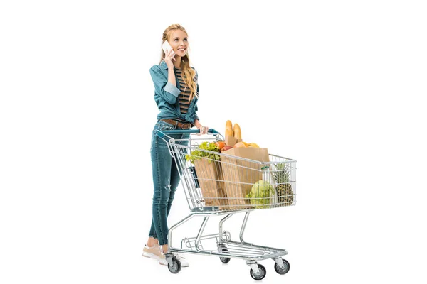 Menina atraente falando no smartphone e transportando carrinho de compras com produtos isolados no branco — Fotografia de Stock