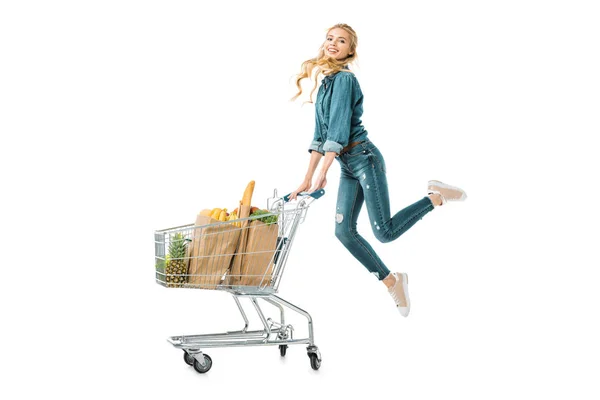 Hermosa chica positiva saltando cerca del carrito de la compra con productos en bolsas de papel aisladas en blanco - foto de stock