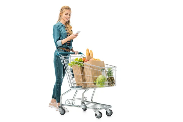 Mujer sonriente usando teléfono inteligente y llevando carrito de compras con productos aislados en blanco - foto de stock