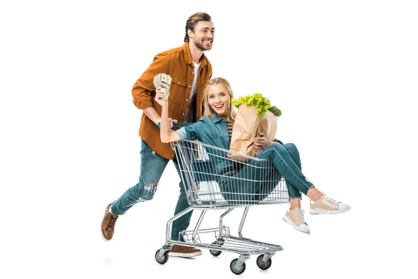 Homme portant chariot d'achat avec petite amie heureuse montrant de l'argent et tenant des sacs en papier avec des produits isolés sur blanc — Photo de stock