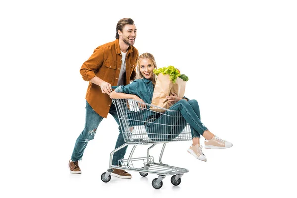 Homem positivo transportando carrinho com namorada feliz segurando sacos de compras com produtos isolados no branco — Fotografia de Stock