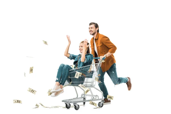 Feliz joven llevando carro con novia mientras ella tirando dinero en efectivo aislado en blanco - foto de stock