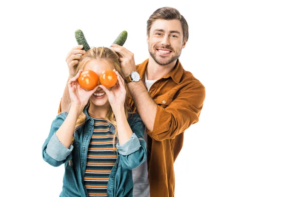 Alegre hombre haciendo cuernos con pepinos a novia mientras ella cubriendo ojos por tomates aislados en blanco - foto de stock