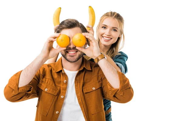 Sorrindo jovem mulher fazendo chifres com bananas para namorado, enquanto ele cobrindo os olhos por laranjas isoladas em branco — Fotografia de Stock