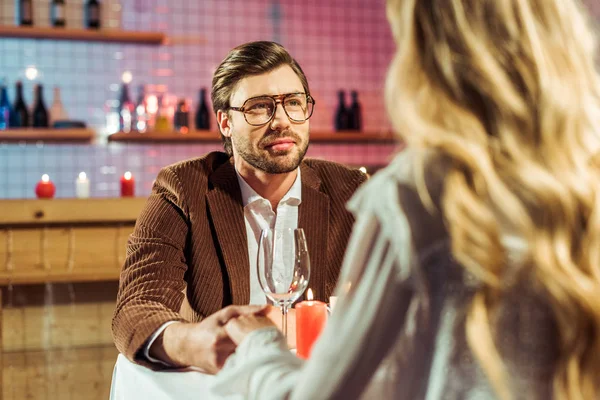Guapo joven en gafas teniendo cita y tomándose de la mano con su novia en el restaurante - foto de stock