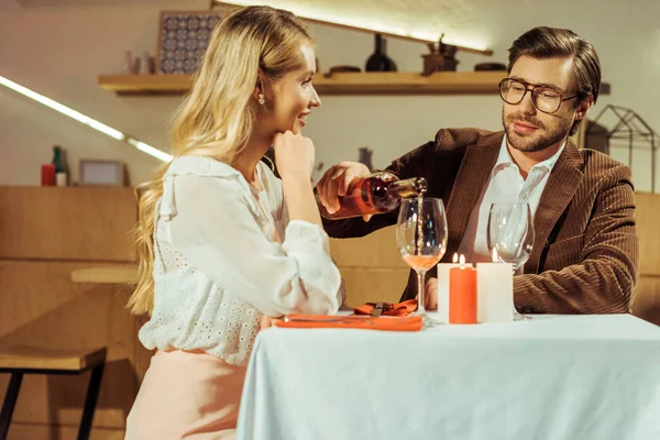 Homem de jaqueta derramando vinho em vidro para bela namorada à mesa no restaurante — Fotografia de Stock