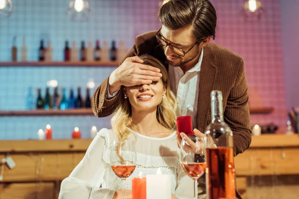 Selektiver Fokus des Mannes in Jacke mit Geschenkbox mit Ring und verdeckenden Augen der Freundin während des romantischen Abendessens im Restaurant — Stockfoto