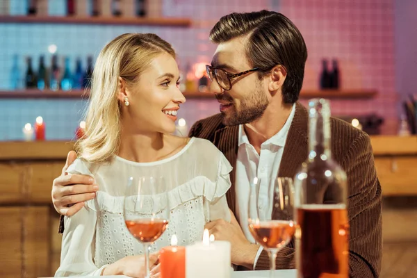 Cheerful man in eyeglasses and jacket embracing girlfriend at table with candles in restaurant — Stock Photo
