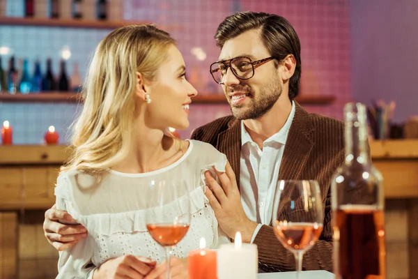 Smiling man in eyeglasses and jacket embracing girlfriend at table with candles in restaurant — Stock Photo