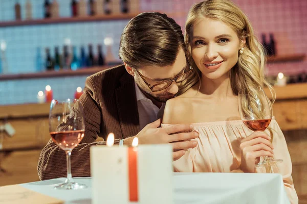 Bela jovem mulher com copo de vinho olhando para a câmera enquanto seu namorado beijando seu ombro na mesa no café — Stock Photo