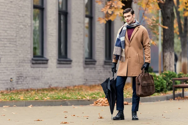 Bell'uomo in cappotto con valigetta e ombrello passeggiando per strada autunnale — Foto stock