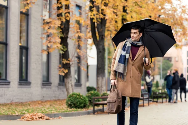 Handsome stylish man in coat and scarf with umbrella walking by autumnal street — Stock Photo