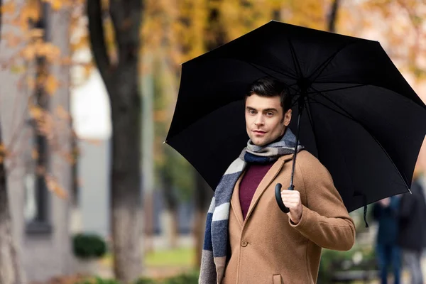 Atractivo hombre elegante en abrigo y bufanda con paraguas caminando por la calle otoñal - foto de stock