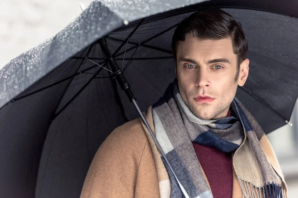 Handsome man in coat and scarf with umbrella looking at camera on street — Stock Photo