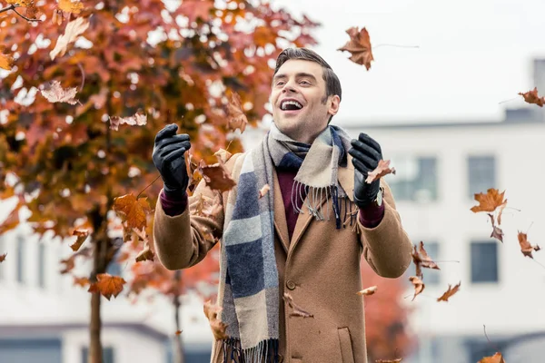 Bel homme heureux en manteau et écharpe vomissant des feuilles tombées dans la rue — Photo de stock