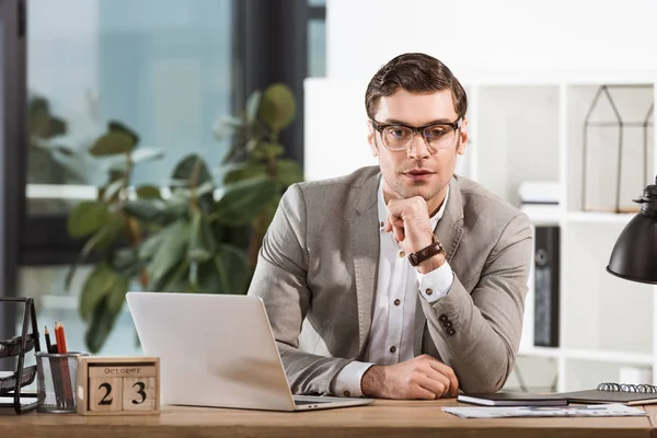 Schöner selbstbewusster Geschäftsmann, der am Arbeitsplatz sitzt und im modernen Büro in die Kamera schaut — Stockfoto