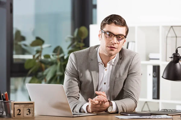 Bell'uomo d'affari di successo seduto sul posto di lavoro e guardando la fotocamera in ufficio moderno — Foto stock