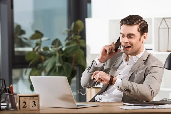 Bell'uomo d'affari felice che parla per telefono sul posto di lavoro in ufficio e guarda lo schermo del portatile — Foto stock