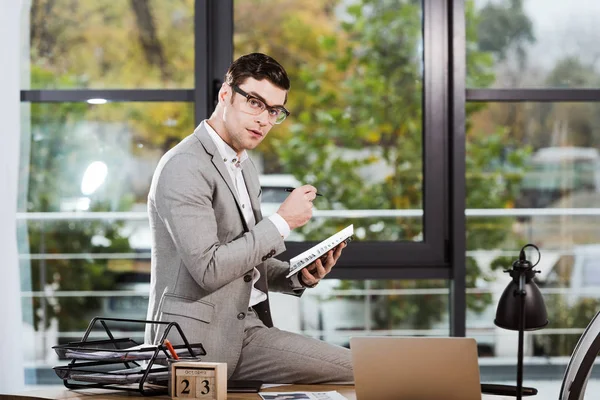 Handsome businessman with notebook sitting at workplace at office and looking at camera — Stock Photo