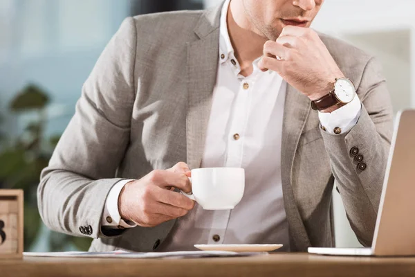 Tiro recortado de homem de negócios com xícara de café olhando para a tela do laptop no escritório — Fotografia de Stock