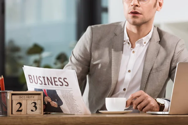 Cropped shot of handsome businessman with cup of coffee reading newspaper at workplace — Stock Photo