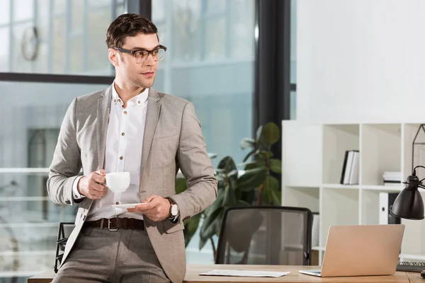 Bell'uomo d'affari con una tazza di caffè appoggiato sul posto di lavoro e distogliendo lo sguardo — Foto stock