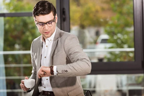 Bel homme d'affaires avec tasse de café en regardant la montre-bracelet au bureau — Photo de stock