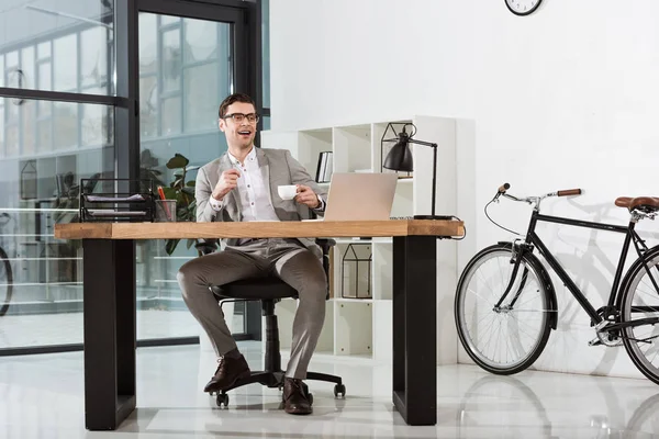 Guapo feliz hombre de negocios con taza de café sentado en el lugar de trabajo en la oficina moderna - foto de stock