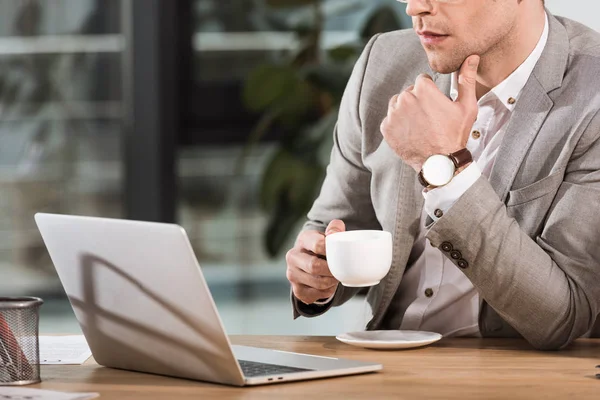 Cropped shot of handsome businessman with cup of coffee using laptop at office — Stock Photo