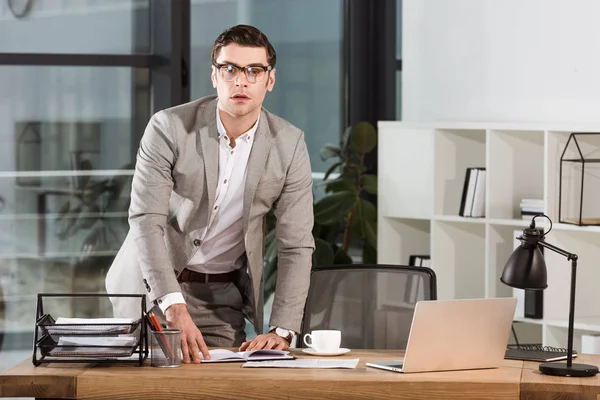 Schöner Geschäftsmann steht am Arbeitsplatz in modernem Büro und blickt in die Kamera — Stockfoto