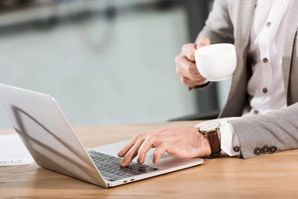 Tiro recortado de homem de negócios com xícara de café usando laptop no escritório — Fotografia de Stock