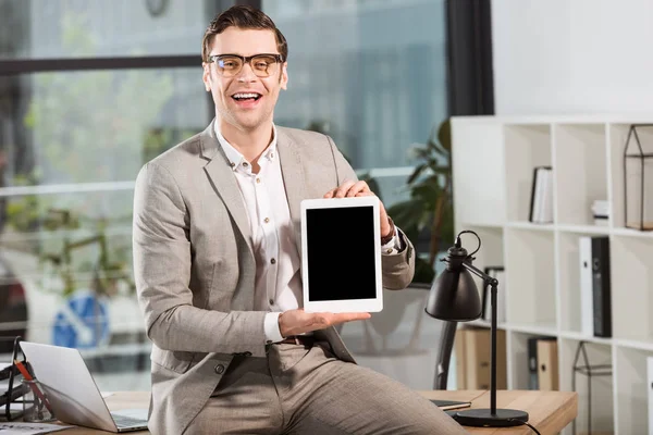 Handsome happy businessman sitting on desk at workplace and holding tablet with blank screen — Stock Photo