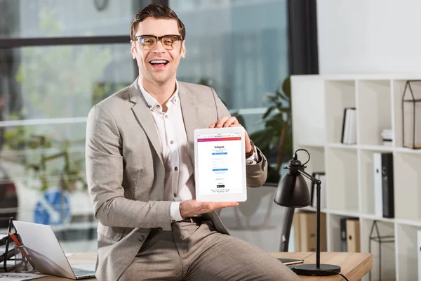 Guapo feliz hombre de negocios sentado en el escritorio en el lugar de trabajo y la celebración de la tableta con Instagram sitio web en la pantalla - foto de stock