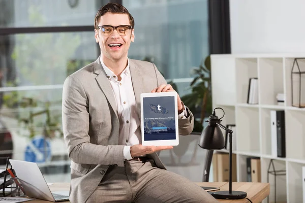 Handsome happy businessman sitting on desk at workplace and holding tablet with tumblr website on screen — Stock Photo