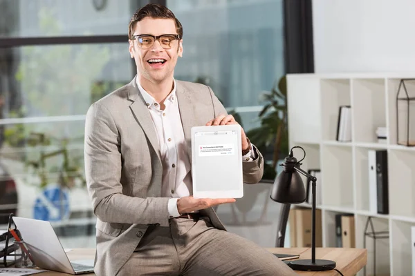 Handsome happy businessman sitting on desk at workplace and holding tablet with blocked vk website on screen — Stock Photo