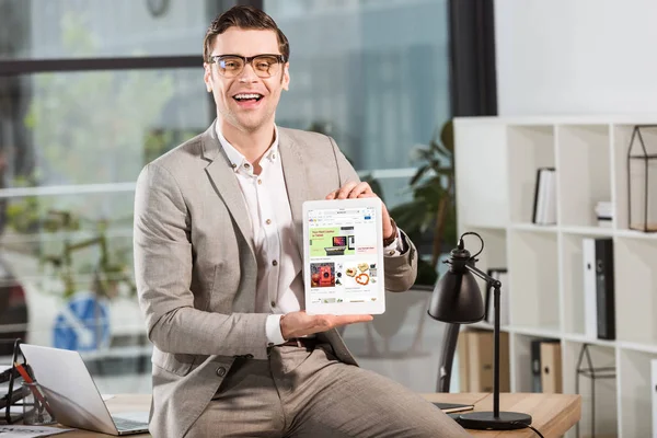 Guapo hombre de negocios feliz sentado en el escritorio en el lugar de trabajo y la celebración de la tableta con ebay sitio web en la pantalla - foto de stock