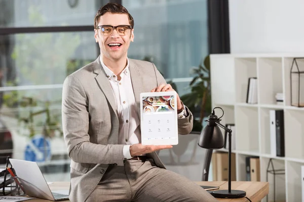 Handsome happy businessman sitting on desk at workplace and holding tablet with foursquare website on screen — Stock Photo