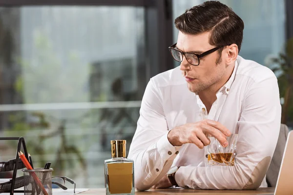 Homme d'affaires déprimé avec un verre de whisky assis sur le lieu de travail au bureau et regardant ailleurs — Photo de stock