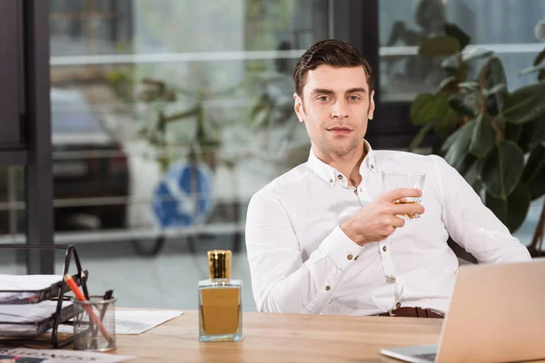 Handsome businessman with glass of whiskey sitting at workplace in office and looking at camera — Stock Photo